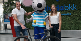 Ein Mann und eine Frau stehen lächelnd neben einer Person in einem großen Mäusemaskottchenkostüm und vor ihnen steht ein schwarzes Fahrrad. Sie befinden sich in einem Gebäude vor einer Wand mit dem Logo der Vivida BKK.