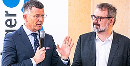 Zwei Männer in Anzügen stehen an einem Tisch mit Mikrofonen und Wasserflaschen. Ein Mann spricht gestikulierend in ein Mikrofon; der andere hört aufmerksam zu. Im Hintergrund ist ein Banner mit einem blauen Logo zu sehen.