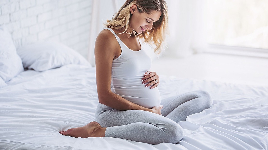 Eine schwangere Frau in einem weißen Tanktop und grauen Leggings sitzt im Schneidersitz auf einem Bett, wiegt sanft ihren Bauch und lächelt in einem hellen, sanft beleuchteten Schlafzimmer.
