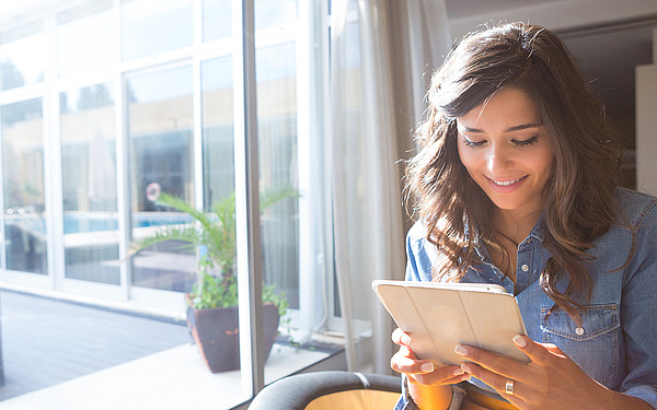 Eine junge Frau mit langen braunen Haaren sitzt lächelnd an einem großen Fenster und benutzt ein Tablet. Sonnenlicht fällt herein, im Hintergrund ist eine Topfpflanze zu sehen.