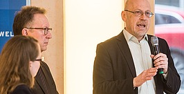 Ein Mann im Anzug spricht in ein Mikrofon und gestikuliert dabei mit der Hand. Zwei weitere Personen, ebenfalls in Business-Kleidung, stehen neben ihm und hören aufmerksam zu. Im Hintergrund sind ein helles Licht und ein rotes Auto zu sehen.