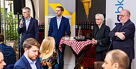 Vier Personen stehen während einer formellen Veranstaltung im vorderen Teil des Raumes und sprechen, während andere an Tischen mit Getränken und Essen sitzen. Im Hintergrund sind ein Banner und Weinflaschen zu sehen.