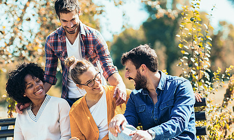 Vier Freunde sitzen an einem sonnigen Tag zusammen auf einer Gartenbank und lächeln und lachen. Zwei Frauen sitzen vor ihnen, eine mit Locken und eine mit Brille, während zwei Männer hinter ihnen stehen und sitzen. Alle wirken glücklich und entspannt.