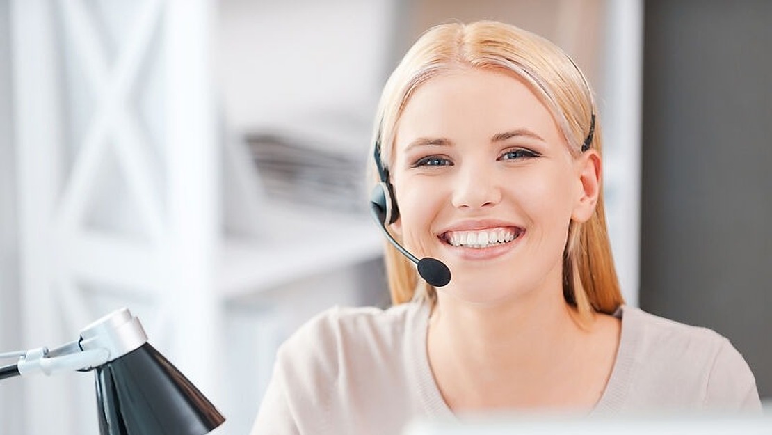 Lächelnde Person mit blonden Haaren, Headset und Sitz an einem Schreibtisch. In der Nähe steht eine schwarz-silberne Schreibtischlampe. Der Hintergrund ist unscharf und suggeriert eine Büroumgebung.