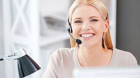 Lächelnde Person mit blonden Haaren, Headset und Sitz an einem Schreibtisch. In der Nähe steht eine schwarz-silberne Schreibtischlampe. Der Hintergrund ist unscharf und suggeriert eine Büroumgebung.
