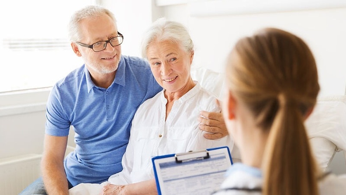 Eine ältere Frau sitzt lächelnd in einem Krankenhausbett. Neben ihr sitzt ein Mann, der ihre Schultern hält. Ihnen gegenüber steht ein Arzt mit einem Klemmbrett in der Hand und spricht mit dem Paar.