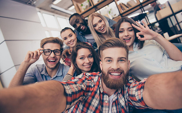 Eine Gruppe von sieben jungen Erwachsenen macht ein fröhliches Selfie in einem Innenbereich und lächelt breit. Sie sitzen zusammen und einige machen Handgesten wie Peace-Zeichen und ein „OK“-Zeichen. Im Hintergrund sind Bücherregale zu sehen.