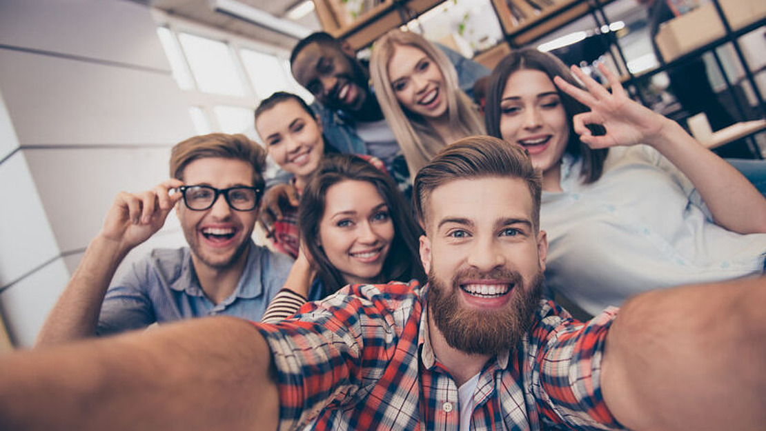 Eine Gruppe von sieben jungen Erwachsenen macht ein fröhliches Selfie in einem Innenbereich und lächelt breit. Sie sitzen zusammen und einige machen Handgesten wie Peace-Zeichen und ein „OK“-Zeichen. Im Hintergrund sind Bücherregale zu sehen.