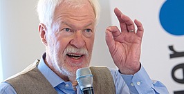 Ein älterer Mann mit weißem Haar und Bart spricht in ein Mikrofon und gestikuliert mit der Hand. Er trägt ein blaues Hemd und eine graue Weste. Im Hintergrund ist ein teilweise sichtbares Logo mit einem blauen Punkt zu sehen.