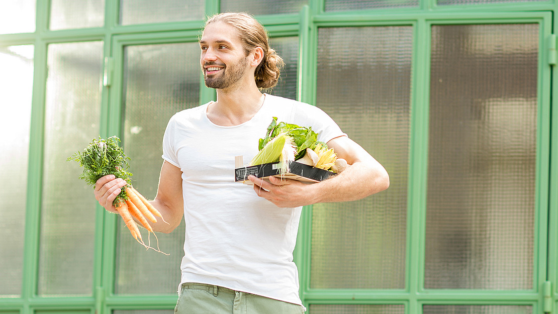 Ein lächelnder Mann mit langem Haar und Pferdeschwanz hält in der einen Hand frische Karotten und in der anderen eine Kiste mit verschiedenem Gemüse und steht vor großen Fenstern mit grünen Rahmen.