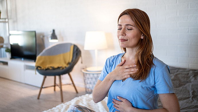 Eine Frau sitzt mit geschlossenen Augen im Haus, eine Hand auf der Brust, die andere auf dem Bauch, und übt in einem ruhigen, gemütlichen Wohnzimmer tiefes Atmen oder Meditation.