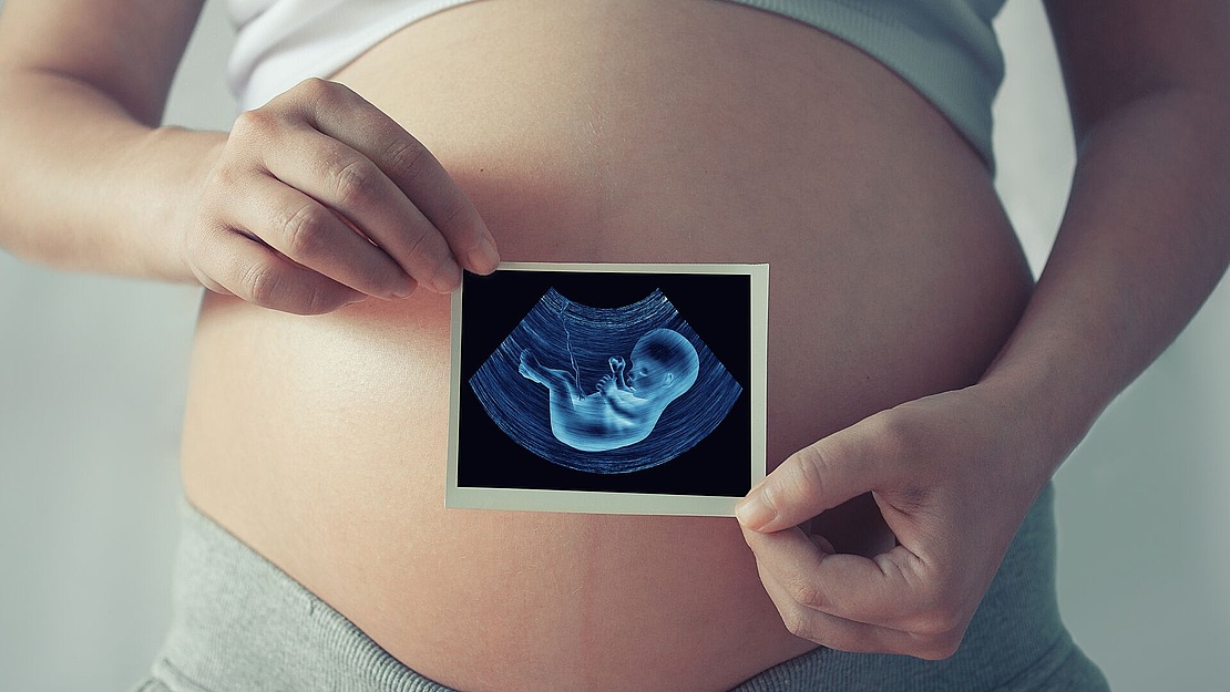 Eine schwangere Person hält ein Ultraschallbild vor ihren nackten Bauch, das einen sich entwickelnden Fötus im Mutterleib zeigt.