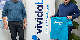 Zwei Männer stehen neben einem „vivida bkk Ihre Krankenkasse“-Banner. Einer hält ein blaues T-Shirt mit weißer Grafik und Text. Sie befinden sich im Innenbereich. Im Hintergrund sind eine Pflanze und ein Plakat zu sehen.