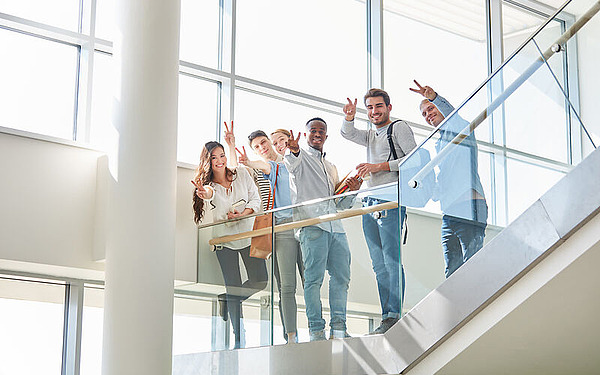 Eine bunt gemischte Gruppe von sechs jungen Erwachsenen steht lächelnd und mit Peace-Zeichen auf einer modernen Treppe in einem hellen, geräumigen Gebäude mit großen Fenstern.