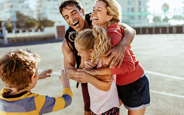 Eine fröhliche vierköpfige Familie, darunter zwei Kinder, umarmt sich und lacht gemeinsam an einem sonnigen Tag auf einem Basketballplatz im Freien.