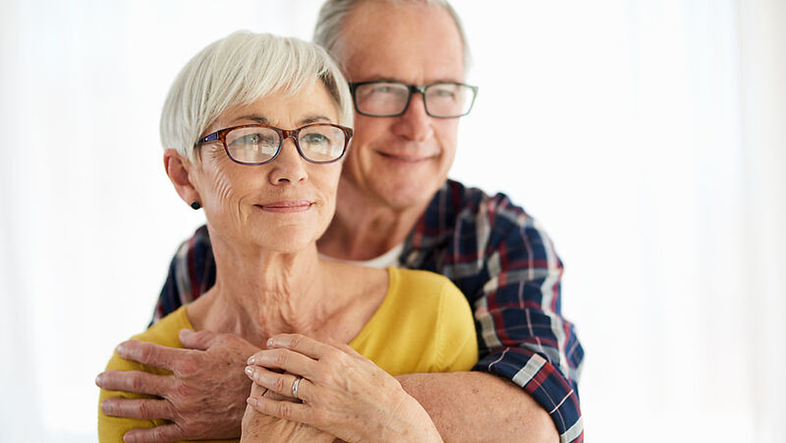 Ein älteres Paar mit Brille steht zusammen und lächelt herzlich. Der Mann umarmt die Frau von hinten, und beide wirken zufrieden. Der Hintergrund ist mit weißen Vorhängen sanft beleuchtet.