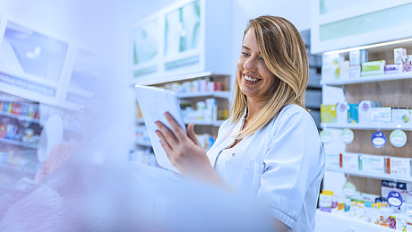 Ein lächelnder Apotheker im weißen Kittel hält ein digitales Tablet in der Hand und betrachtet es, während er in einer hell erleuchteten Apotheke steht, im Hintergrund stehen Regale voller Medikamente und Gesundheitsprodukte.