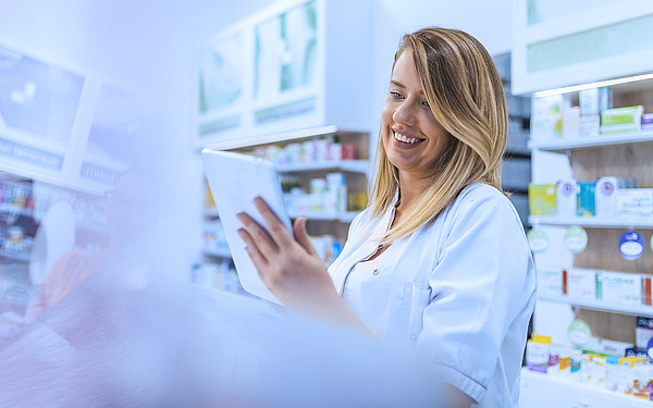 Ein lächelnder Apotheker im weißen Kittel hält ein digitales Tablet in der Hand und betrachtet es, während er in einer hell erleuchteten Apotheke steht, im Hintergrund stehen Regale voller Medikamente und Gesundheitsprodukte.