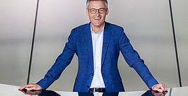 Ein Mann mittleren Alters mit Brille und blauer Anzugsjacke steht hinter einem glänzenden Schreibtisch und lächelt in die Kamera vor einem hellgrauen Hintergrund mit dunklen Linien.