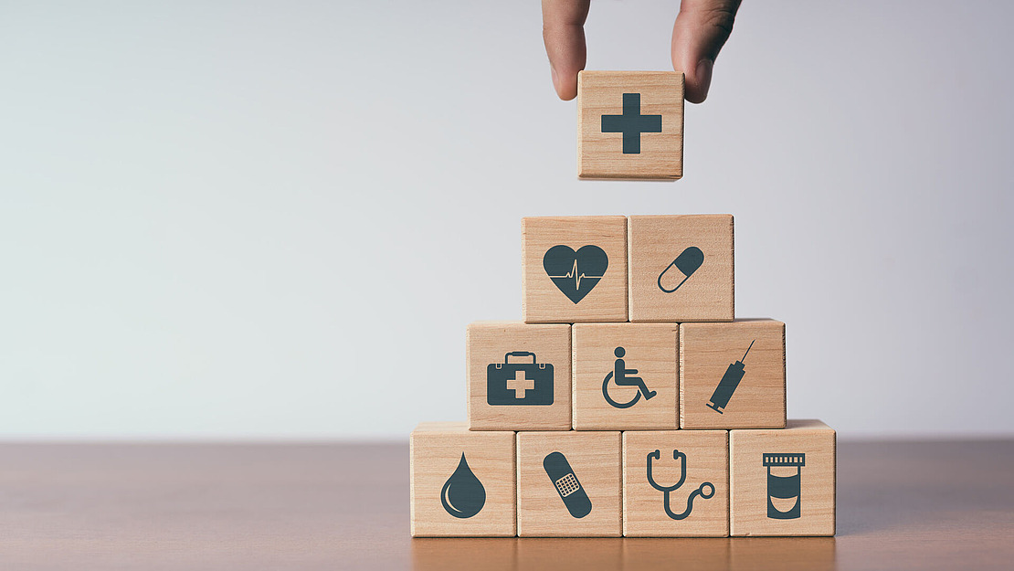 Eine Hand legt einen Holzblock mit einem medizinischen Kreuz auf eine Pyramide aus Blöcken, von denen jeder unterschiedliche Gesundheitssymbole zeigt, beispielsweise einen Herzmonitor, eine Spritze, eine Pille, einen Rollstuhl, einen Tropfen, einen Verband, ein Stethoskop und eine Medikamentenflasche.
