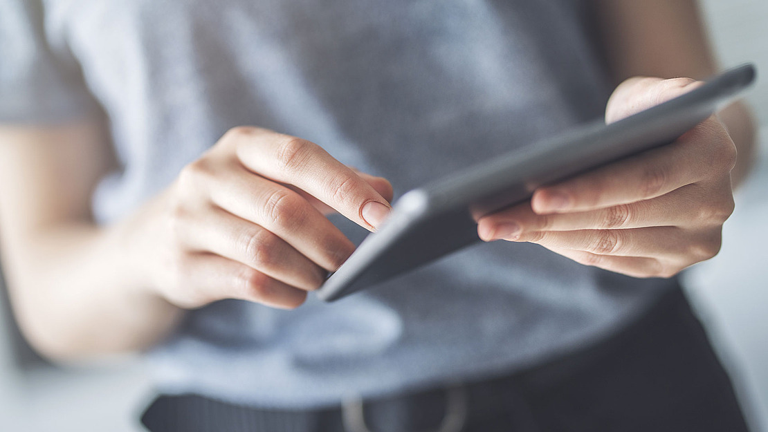 Eine Person in einem grauen Hemd hält ein Tablet in der einen Hand und berührt mit der anderen den Bildschirm. Der Hintergrund ist unscharf.