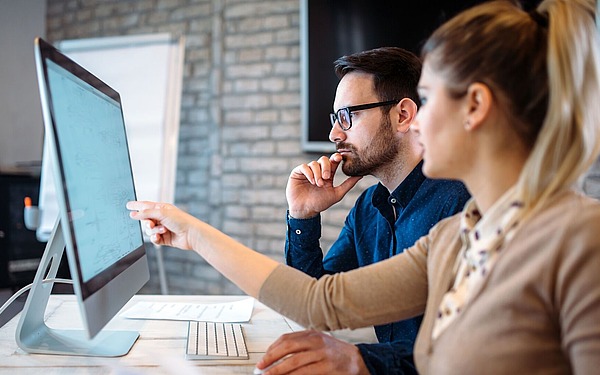 Zwei Personen sitzen an einem Schreibtisch und schauen konzentriert auf einen Computermonitor. Die Frau zeigt auf den Bildschirm, während der Mann sein Kinn auf seine Hand stützt. Beide scheinen konzentriert und in eine Diskussion vertieft zu sein.