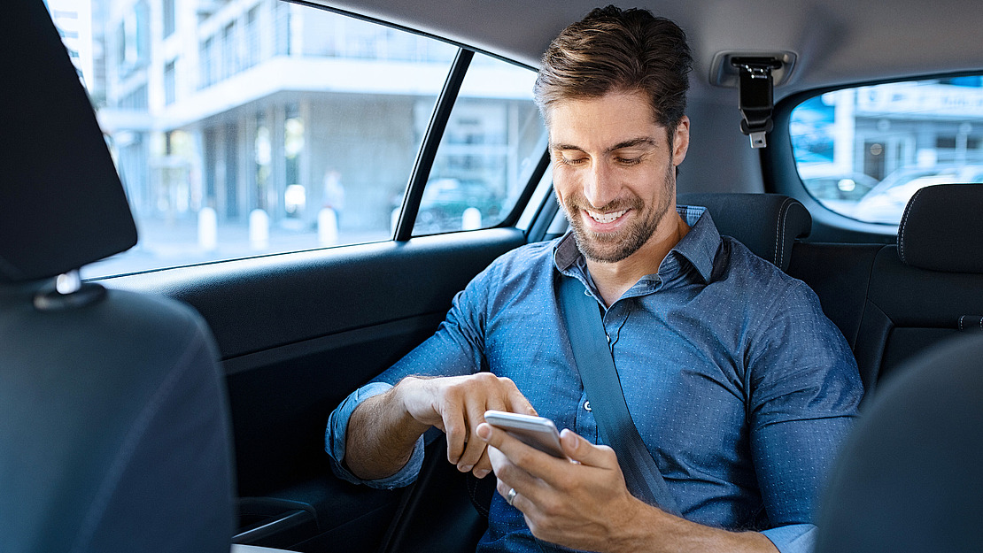 Ein Mann in einem blauen Hemd sitzt auf dem Rücksitz eines Autos, lächelt und benutzt sein Smartphone. Er ist angeschnallt, und aus dem Fenster sind städtische Gebäude zu sehen.