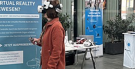 Eine Frau, die ein VR-Headset trägt und einen Controller in der Hand hält, steht vor einem Informationsbanner über ein Virtual-Reality-Modell des Dickdarms.
