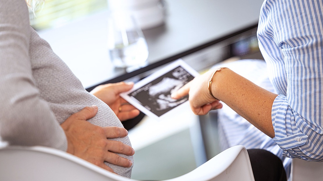 Eine schwangere Person hält ihren Bauch, während eine andere Person in einem gestreiften Hemd auf ein Ultraschallbild zeigt, beide sitzen an einem Tisch mit einem Glas Wasser und Papieren im Hintergrund.