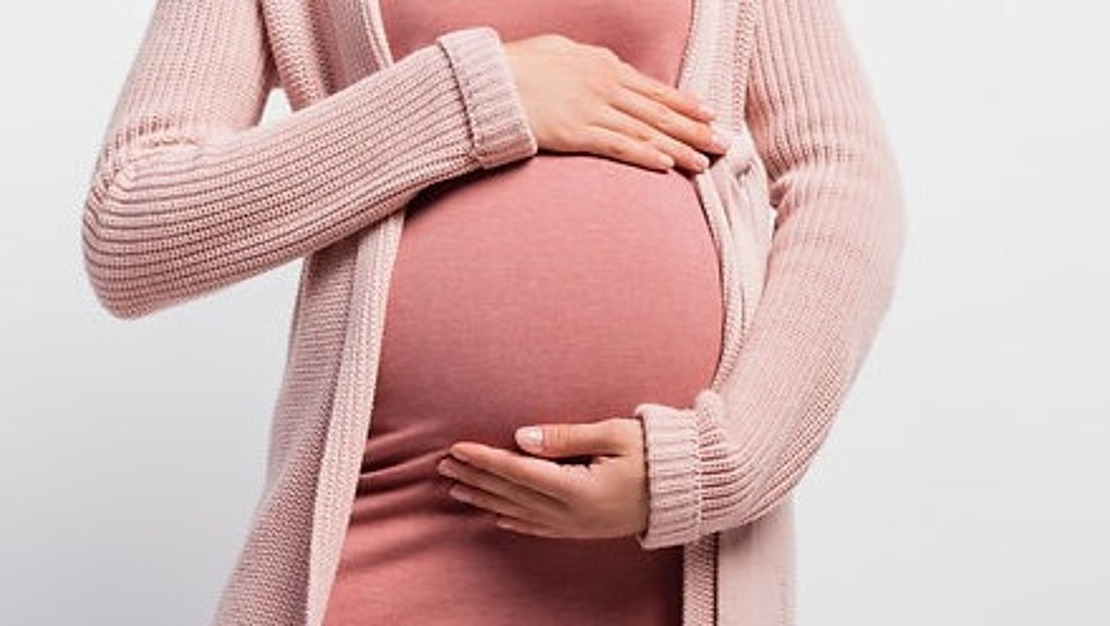 Eine schwangere Person trägt ein rosa Kleid und eine Strickjacke und hält ihren Bauch sanft mit beiden Händen. Der Hintergrund ist schlicht weiß.