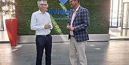 Zwei Männer stehen in einer modernen Lobby mit einer grünen Pflanzenwand, auf der das Wort „vivida“ zu lesen ist, und unterhalten sich. Der eine trägt ein weißes Hemd und eine dunkle Hose, der andere eine karierte Jacke, ein rosa Hemd und eine Khakihose.