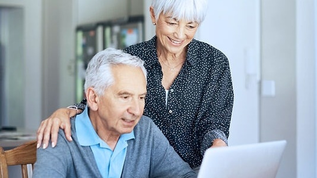Ein älteres Ehepaar sitzt an einem Esstisch und benutzt einen Laptop. Der Mann sitzt und tippt, während die Frau neben ihm steht, lächelt und auf den Bildschirm schaut. Auf dem Tisch stehen auch eine Kaffeetasse und Papiere.
