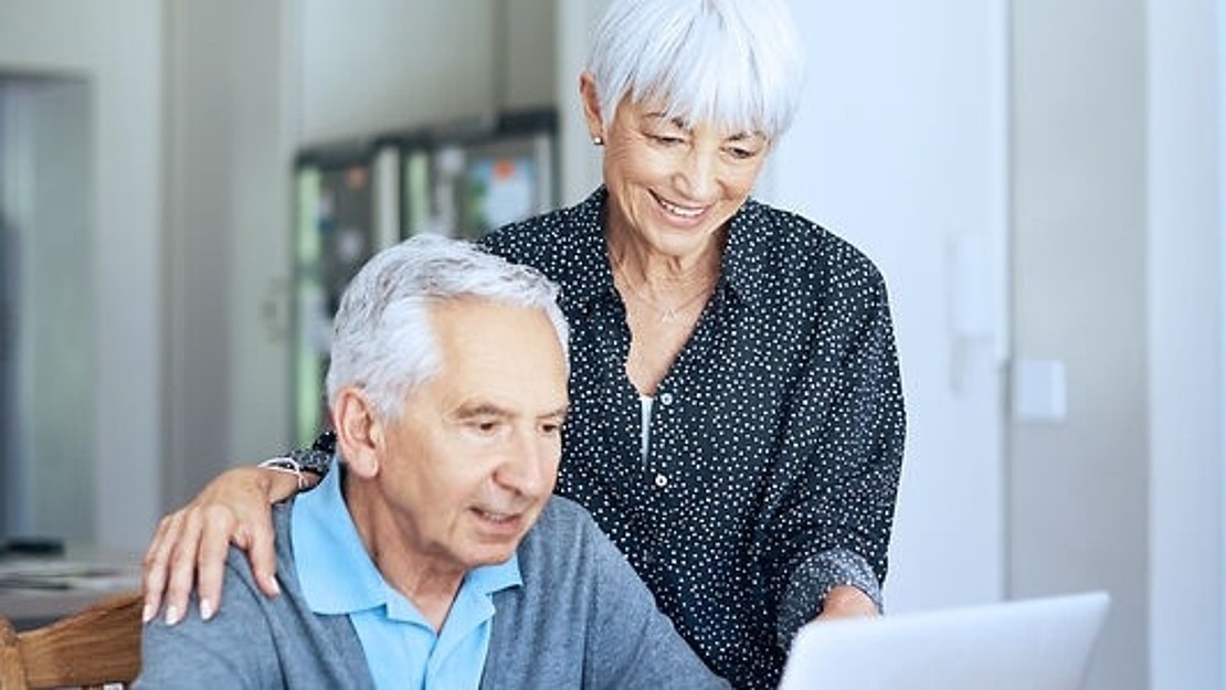 Ein älteres Ehepaar sitzt an einem Esstisch und benutzt einen Laptop. Der Mann sitzt und tippt, während die Frau neben ihm steht, lächelt und auf den Bildschirm schaut. Auf dem Tisch stehen auch eine Kaffeetasse und Papiere.
