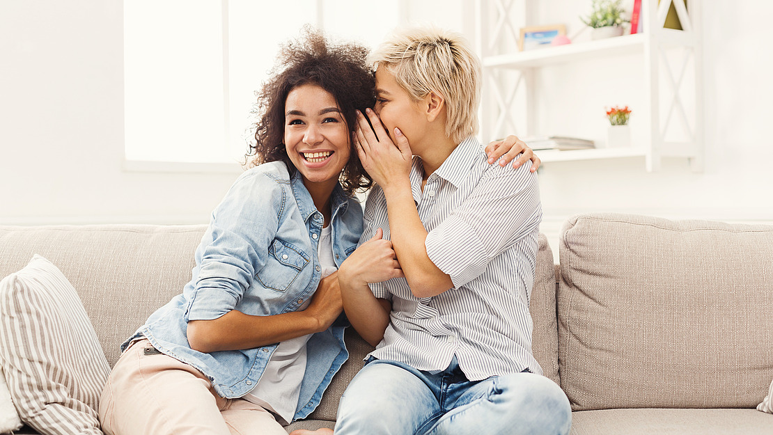 Zwei Menschen sitzen eng beieinander auf einem Sofa. Einer lächelt breit in die Kamera, während der andere sich vorbeugt und ihm etwas ins Ohr flüstert. Beide wirken glücklich und entspannt in einem hellen, gemütlichen Wohnzimmer.