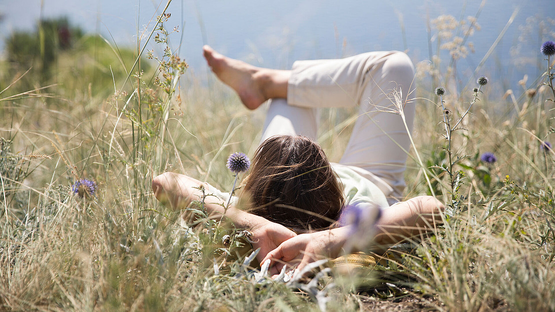 Eine Person liegt auf dem Rücken in hohem Gras und Wildblumen, die Arme hinter dem Kopf verschränkt und die Beine angewinkelt, und entspannt sich im Freien unter einem strahlenden Himmel. Die Szene vermittelt ein Gefühl von Ruhe und Entspannung in der Natur.