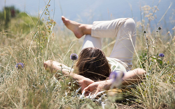 Eine Person liegt auf dem Rücken in hohem Gras und Wildblumen, die Arme hinter dem Kopf verschränkt und die Beine angewinkelt, und entspannt sich im Freien unter einem strahlenden Himmel. Die Szene vermittelt ein Gefühl von Ruhe und Entspannung in der Natur.