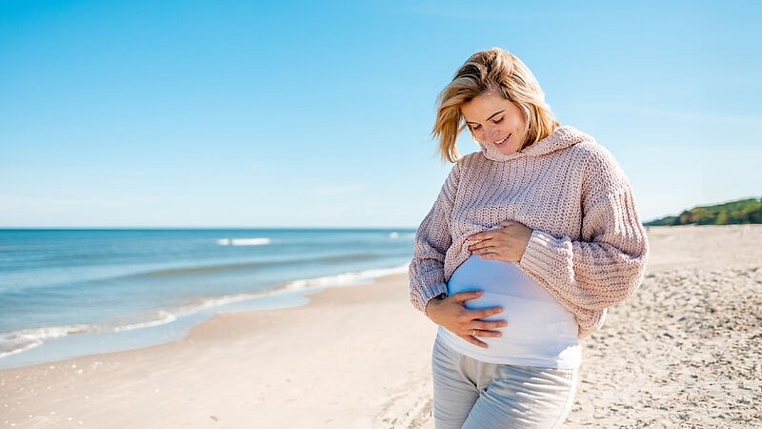 Eine schwangere Frau in einem rosa Pullover und einer weißen Hose steht lächelnd an einem Sandstrand und hält sanft ihren Bauch. Im Hintergrund ist das Meer unter einem klaren blauen Himmel ruhig.