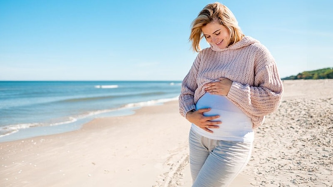 Eine schwangere Frau in einem rosa Pullover und einer weißen Hose steht lächelnd an einem Sandstrand und hält sanft ihren Bauch. Im Hintergrund ist das Meer unter einem klaren blauen Himmel ruhig.