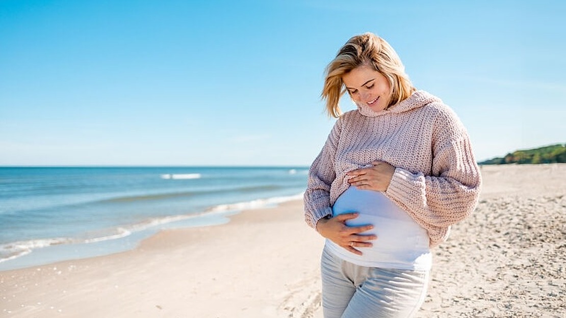 Eine schwangere Frau in einem rosa Pullover und einer weißen Hose steht lächelnd an einem Sandstrand und hält sanft ihren Bauch. Im Hintergrund ist das Meer unter einem klaren blauen Himmel ruhig.
