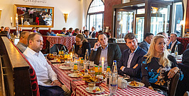 Eine Gruppe von Personen in Businesskleidung sitzt an einem Restauranttisch mit karierter Tischdecke und hört aufmerksam zu. Teller mit Essen, Getränken und Kerzen stehen auf dem Tisch. Im Hintergrund ist das Restaurant gut besucht.