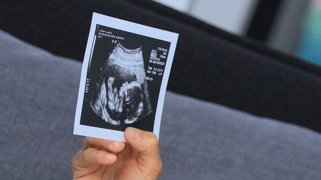 Eine Hand hält ein Schwarz-Weiß-Ultraschallfoto, das wahrscheinlich ein sich entwickelndes Baby zeigt, vor einem unscharfen Hintergrund hoch.