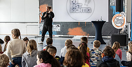 Ein Mann spricht auf der Bühne zu einer Gruppe sitzender Kinder in einer Turnhalle. Hinter ihm ist eine Leinwand mit der Aufschrift „DU BIST RICHTIG“ und bunten Verzierungen zu sehen. Die Kinder schauen aufmerksam zu.