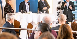 Vier Personen stehen und sitzen um einen hohen Tisch auf einer Bühne und diskutieren vor Publikum. Ein Mann spricht in ein Mikrofon. Auf den Transparenten sind unter anderem die Aufschriften „Deutsche Welle“ und „Schwenninger“ zu sehen.