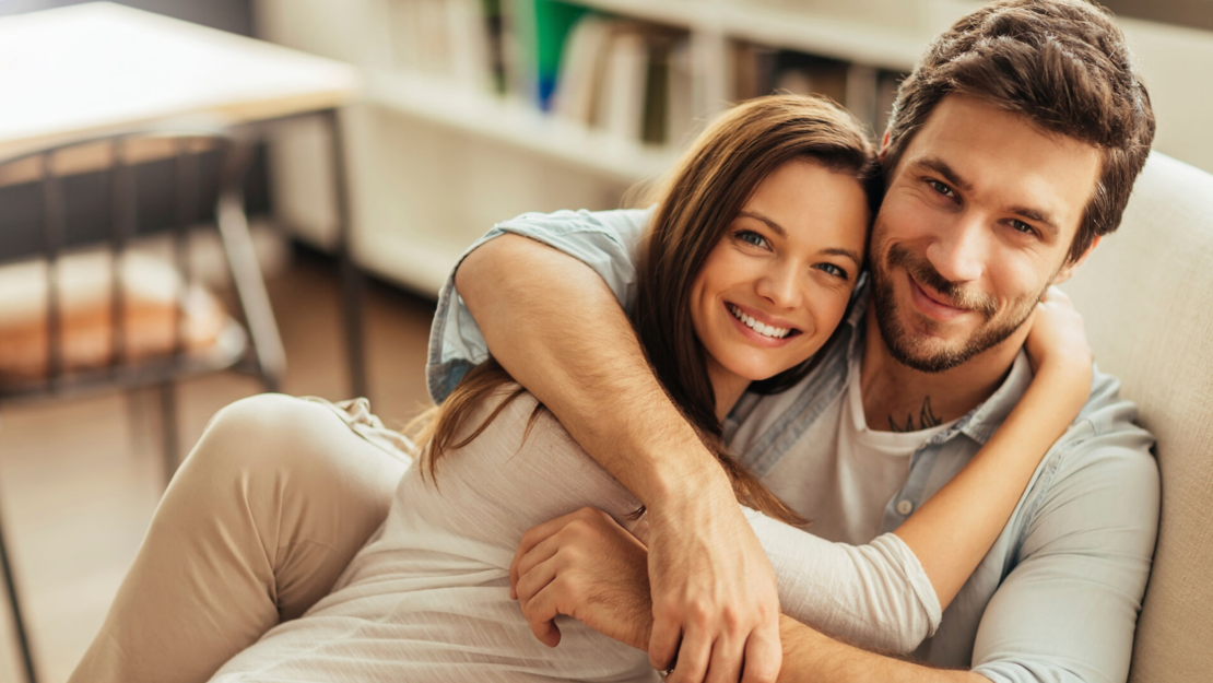 Ein lächelndes Paar sitzt eng aneinander geschmiegt auf einer Couch und umarmt sich gegenseitig. Beide haben braunes Haar und wirken glücklich und entspannt in einem gemütlichen, gut beleuchteten Raum mit Bücherregalen im Hintergrund.
