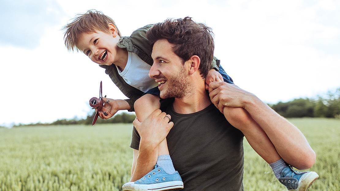 Ein lächelnder Mann trägt ein glückliches Kind auf seinen Schultern auf einer üppigen grünen Wiese. Das Kind hält ein kleines Spielzeugflugzeug. Beide blicken in die Ferne und genießen einen sonnigen Tag im Freien.