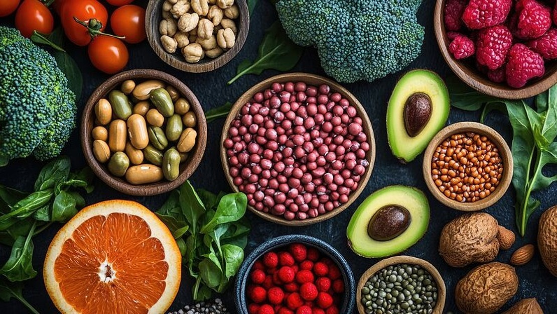 Eine Auswahl an frischem Obst, Gemüse, Hülsenfrüchten, Nüssen und Samen, die auf einer dunklen Oberfläche angeordnet sind; enthält Brokkoli, Avocado, Tomaten, Orangen, Himbeeren, Bohnen, Walnüsse und verschiedene Samen in kleinen Schalen.