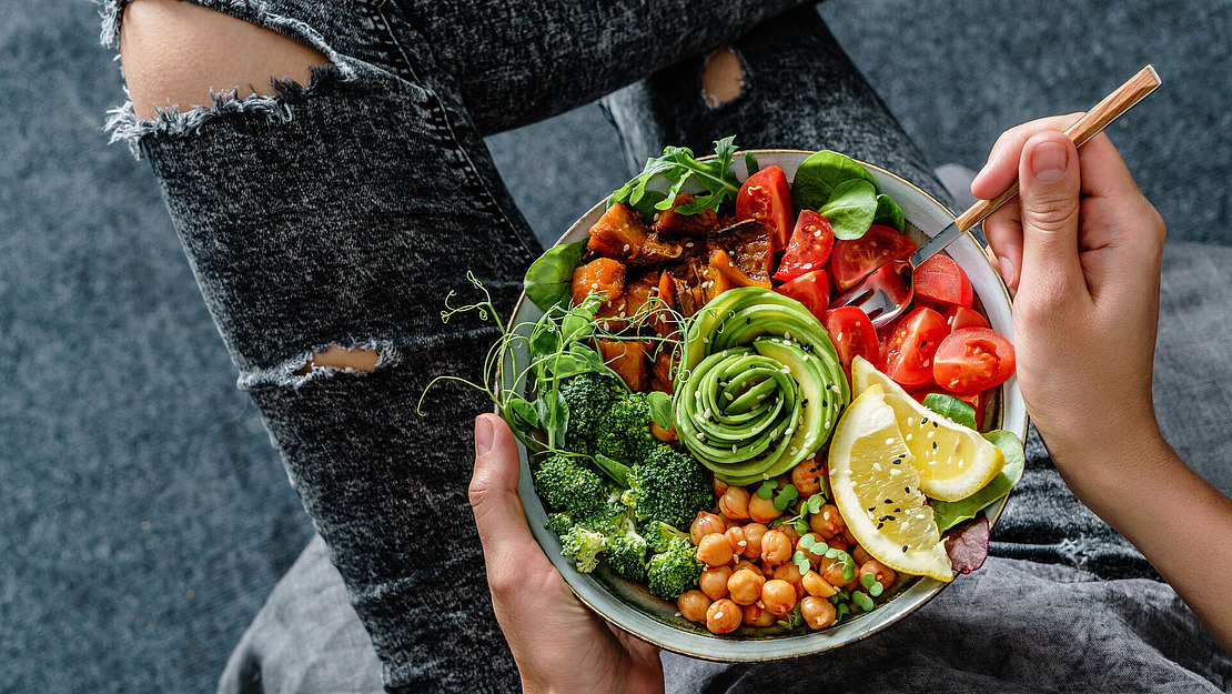 Eine Person in zerrissenen Jeans hält eine bunte Schüssel gefüllt mit Avocado-Spiralen, Brokkoli, Kichererbsen, Erbsen, gerösteten Süßkartoffeln, Kirschtomaten, Zitronenscheiben und Blattgemüse und isst mit einer Gabel.