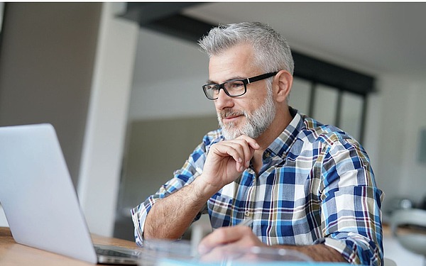 Ein Mann mittleren Alters mit grauem Haar und Bart, der eine Brille und ein kariertes Hemd trägt, sitzt an einem Tisch und schaut nachdenklich auf einen Laptop-Bildschirm in einem hellen, modernen Haus.