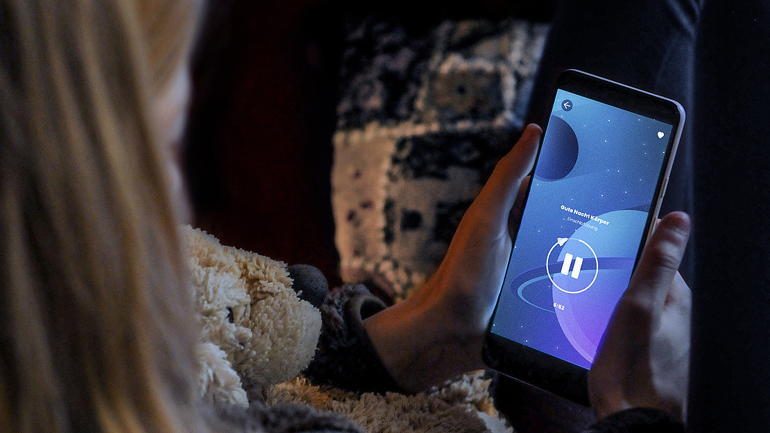 Eine Person sitzt mit einem Teddybären in der Hand da und blickt auf einen Smartphone-Bildschirm mit pausiertem Audiotrack und Weltraum-Hintergrund. Die Szene wirkt gemütlich und entspannt.
