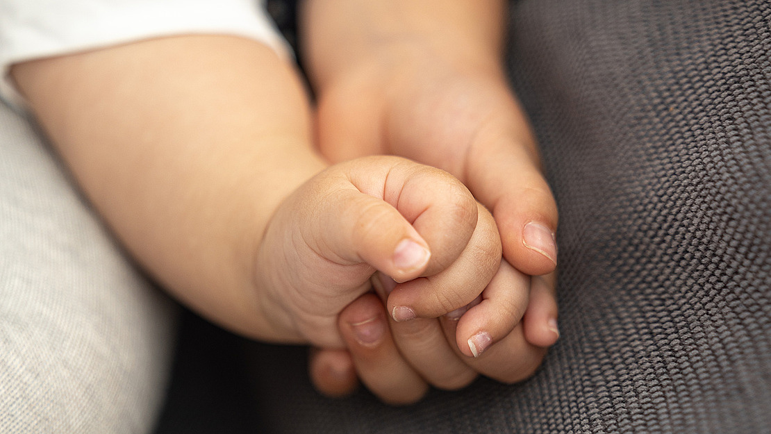 Nahaufnahme einer Babyhand, die sanft eine Erwachsenenhand auf einer strukturierten grauen Stoffoberfläche hält und so Verbindung und Fürsorge symbolisiert.